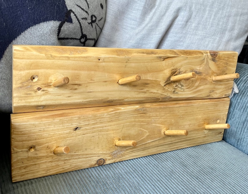 May include: A wooden wall-mounted coat rack with six wooden pegs. The rack is made of two pieces of reclaimed wood, stained in a light brown colour.