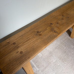 May include: A long, rectangular wooden bench with a natural finish. The bench has a light brown color with visible wood grain and knots. The legs are also made of wood and are attached to the underside of the bench.