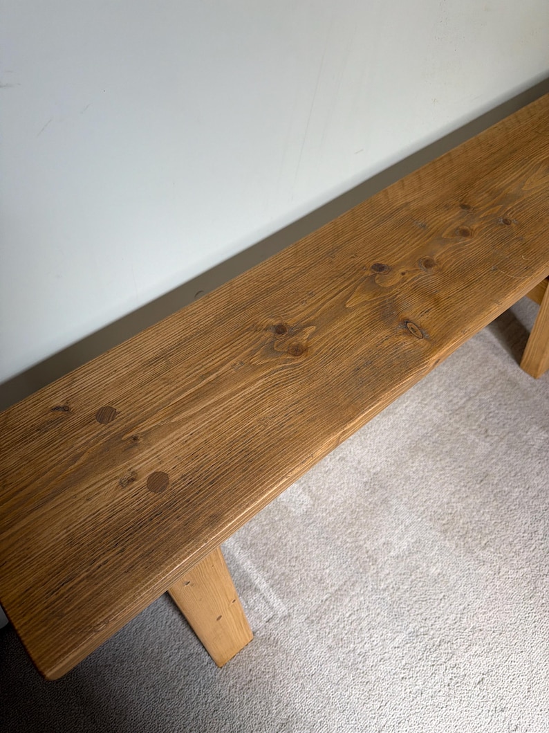 May include: A long, rectangular wooden bench with a natural finish. The bench has a light brown color with visible wood grain and knots. The legs are also made of wood and are attached to the underside of the bench.