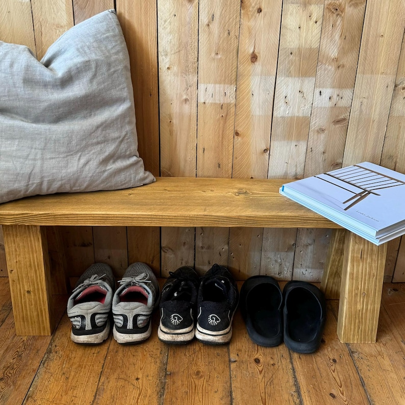 May include: A wooden bench with a grey cushion on top. The bench has a shelf underneath, where there are four pairs of shoes and a book.