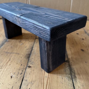 May include: A dark grey wooden stool with a rectangular top and two legs. The stool is made of rough-hewn wood and has a rustic appearance.