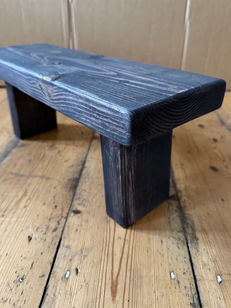 May include: A dark grey wooden stool with a rectangular top and two legs. The stool is made of rough-hewn wood and has a rustic appearance.