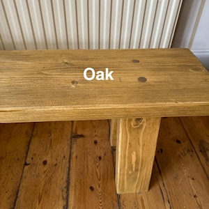 May include: A light brown wooden bench with the word "Oak" printed on the top. The bench has a rectangular top and four sturdy legs. The bench is made of wood and has a natural wood grain. The bench is suitable for indoor use.