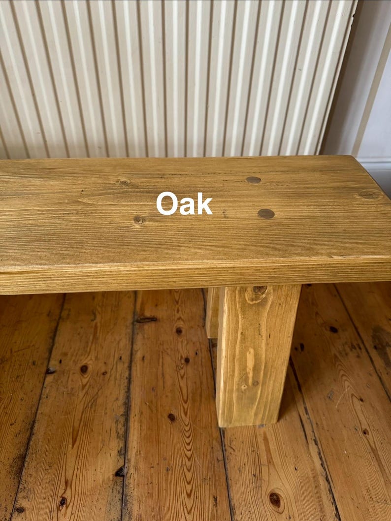 May include: A light brown wooden bench with the word "Oak" printed on the top. The bench has a rectangular top and four sturdy legs. The bench is made of wood and has a natural wood grain. The bench is suitable for indoor use.