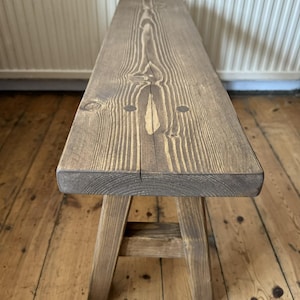 May include: A rustic wooden bench with a natural finish. The bench features a rectangular seat supported by four angled legs. The wood grain is visible, and the bench has a weathered appearance, suggesting it is handmade. The bench is suitable for indoor or outdoor use.