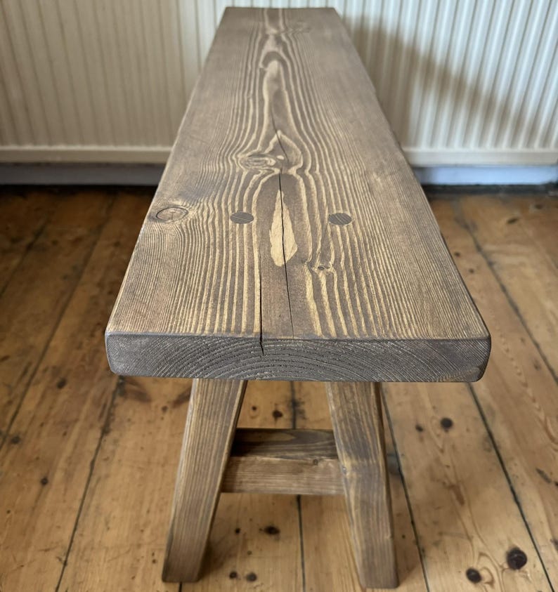 May include: A rustic wooden bench with a natural finish. The bench features a rectangular seat supported by four angled legs. The wood grain is visible, and the bench has a weathered appearance, suggesting it is handmade. The bench is suitable for indoor or outdoor use.