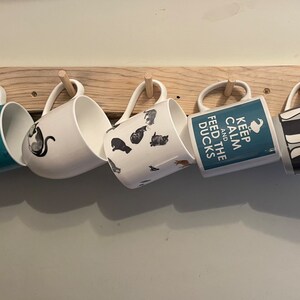 May include: A wooden mug rack with five mugs hanging from it. The mugs have various designs, including a black and white cat, a blue mug with a black and white dog, a white mug with black and white cats, a blue mug with the text "Keep Calm and Feed the Ducks", and a grey mug with black and white penguins.