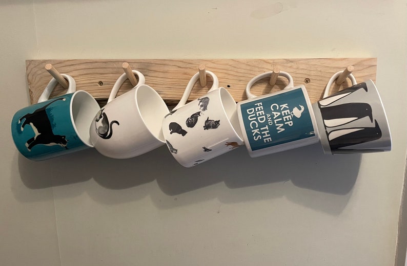 May include: A wooden mug rack with five mugs hanging from it. The mugs have various designs, including a black and white cat, a blue mug with a black and white dog, a white mug with black and white cats, a blue mug with the text "Keep Calm and Feed the Ducks", and a grey mug with black and white penguins.
