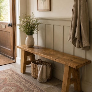 May include: A rustic wooden bench with angled legs sits in a hallway. A woven basket with a fringed throw is beneath it. A vase of flowers and a framed botanical print add to the room's decor. A coat hangs on a wall hook.