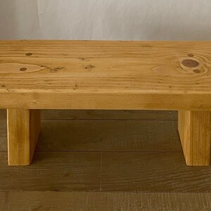 May include: A simple wooden bench with a natural finish. The bench has a rectangular top and two square legs. The bench is made of solid wood and has a rustic look.