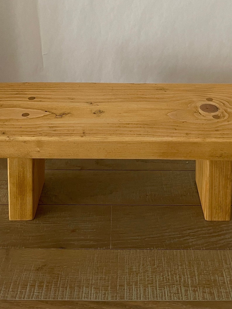 May include: A simple wooden bench with a natural finish. The bench has a rectangular top and two square legs. The bench is made of solid wood and has a rustic look.