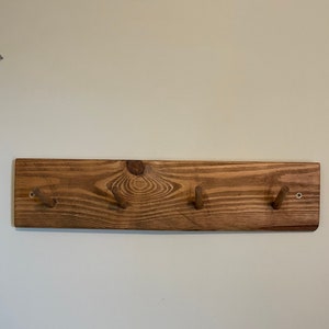 May include: A wooden wall-mounted coat rack with four hooks. The rack is made of natural wood with a dark brown stain. The hooks are made of wood and are attached to the rack with screws.