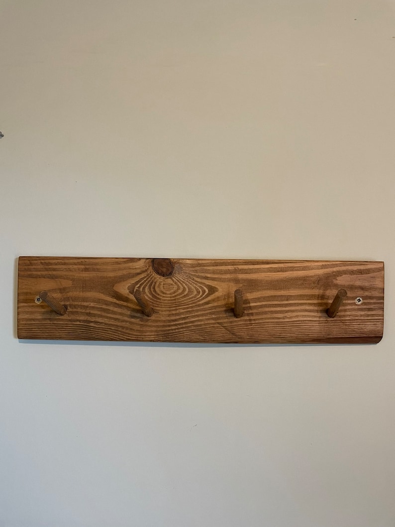 May include: A wooden wall-mounted coat rack with four hooks. The rack is made of natural wood with a dark brown stain. The hooks are made of wood and are attached to the rack with screws.