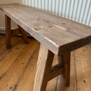 May include: A rustic wooden bench with a rectangular seat and angled legs. The bench is made of stained wood, showcasing the natural grain and knots. The bench is a warm brown color and appears to be handmade.