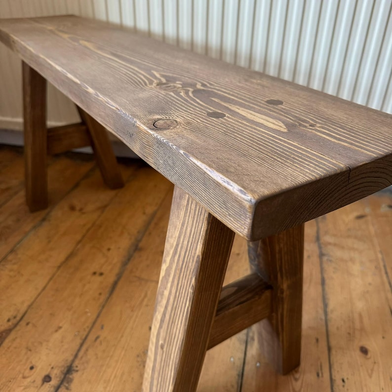 May include: A rustic wooden bench with a rectangular seat and angled legs. The bench is made of stained wood, showcasing the natural grain and knots. The bench is a warm brown color and appears to be handmade.