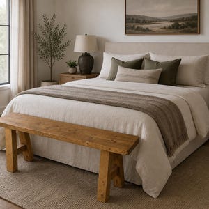 May include: A bedroom scene featuring a bed with cream-colored bedding, olive green pillows, and a brown throw blanket. A wooden bench sits at the foot of the bed. A landscape painting hangs above the bed.