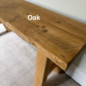 May include: A wooden bench with a natural oak finish. The bench has a rectangular top and angled legs. The wood grain is visible, and the word "Oak" is printed on the top. The bench is suitable for indoor or outdoor use.