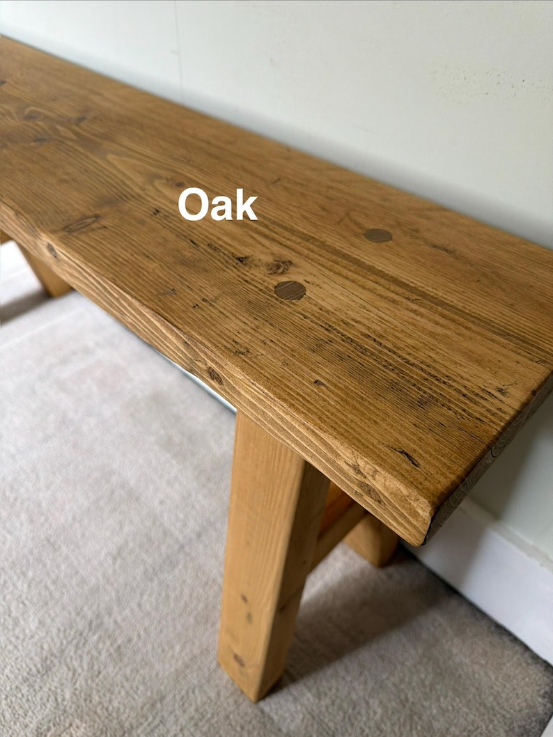 May include: A wooden bench with a natural oak finish. The bench has a rectangular top and angled legs. The wood grain is visible, and the word "Oak" is printed on the top. The bench is suitable for indoor or outdoor use.