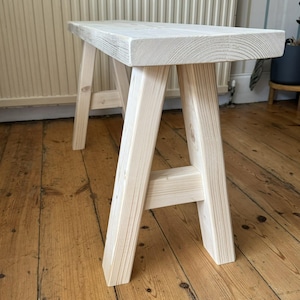 May include: A light-colored wooden bench with a rectangular seat and A-frame legs. The bench is made of unfinished wood, showcasing the natural grain. The bench measures approximately 36 inches long, 12 inches wide, and 18 inches high.