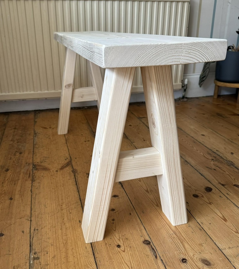 May include: A light-colored wooden bench with a rectangular seat and A-frame legs. The bench is made of unfinished wood, showcasing the natural grain. The bench measures approximately 36 inches long, 12 inches wide, and 18 inches high.