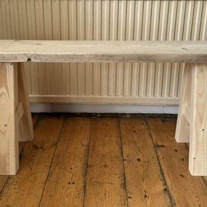 May include: A light-colored wooden bench with a rectangular top and four sturdy legs. The bench is made of unfinished wood, showcasing the natural grain and texture. The bench is approximately 48 inches long.