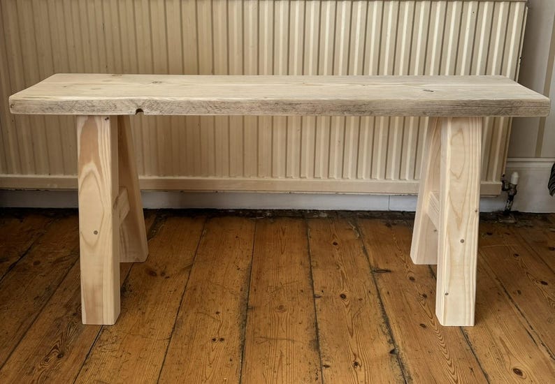 May include: A light-colored wooden bench with a rectangular top and four sturdy legs. The bench is made of unfinished wood, showcasing the natural grain and texture. The bench is approximately 48 inches long.