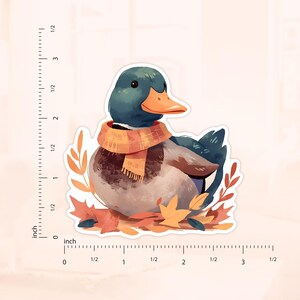May include: A sticker featuring a watercolor illustration of a duck wearing an orange scarf, surrounded by autumn leaves. The duck has a teal head and a bright orange beak. The sticker measures approximately 3 inches in length.