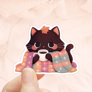 May include: A sticker featuring a cartoon black cat with a fall leaf on its head, wrapped in a blanket and holding a mug of coffee. The blanket is patterned with stars and geometric shapes in shades of pink, orange, and blue.