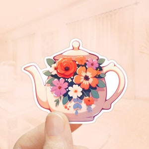 Pode incluir: Uma chaleira branca com um design floral. A chaleira está cheia de flores coloridas, incluindo laranja, rosa e amarelo. As flores estão dispostas em forma de buquê.