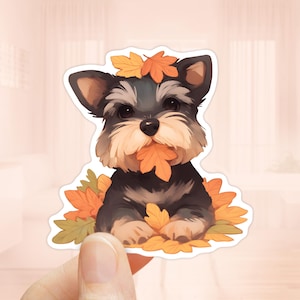 Puede incluir: Una pegatina con un perro de dibujos animados con pelaje negro y gris, rodeado y sosteniendo hojas de otoño. El perro tiene una expresión dulce y está sentado entre las coloridas hojas, creando un tema otoñal.