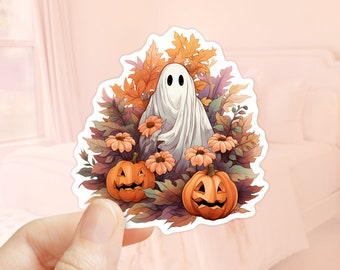 Peut inclure: Un autocollant représentant un fantôme de dessin animé avec un drap blanc, entouré de citrouilles oranges, de fleurs et de feuilles d'automne. Le fantôme a deux yeux noirs. L'autocollant est tenu par une main.