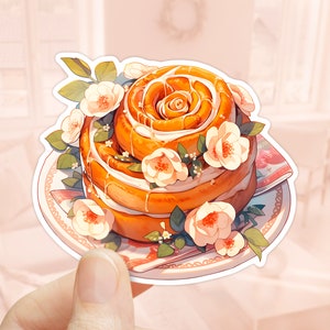 Könnte beinhalten: Eine Zimtschnecke in Form einer Rose, verziert mit weißem Zuckerguss und weißen Blüten. Die Schnecke liegt auf einem weißen Teller mit floralem Muster.
