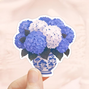 Könnte beinhalten: Ein Aufkleber mit einem Strauß blauer und weißer Hortensien in einer blau-weiß gemusterten Vase. Der Aufkleber hat einen weißen Rand und wird von einer Hand gehalten.