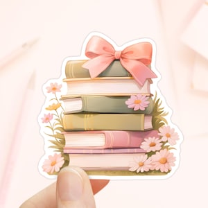 May include: A stack of books with a pink bow on top, surrounded by pink flowers. The books are various shades of brown and green, with a pink and white book on top.