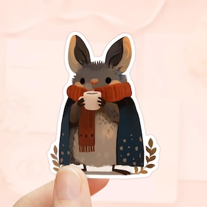 May include: A sticker of a cartoon bat-like creature with large ears, wearing a red scarf and a blue cape with white dots. The creature holds a white mug with a brown liquid, and is surrounded by brown leaves.