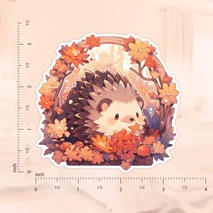 May include: A sticker featuring a cute hedgehog illustration. The hedgehog is surrounded by autumn leaves and flowers in shades of orange, yellow, and brown. The sticker has a white border and is approximately 7.6 cm wide.