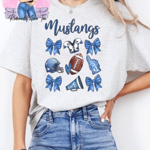 May include: A gray t-shirt with the word "Mustangs" printed on it. The shirt has a variety of blue and white cheerleading and football-themed graphics, including bows, a football helmet, a football, a cheerleader's pom-pom, and a megaphone.