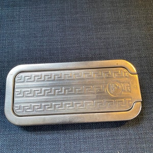 Puede incluir: Una caja rectangular de metal plateado con un patrón de llave griega y un pequeño logotipo en la parte superior. La caja tiene una tapa con bisagras.