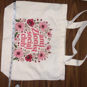 K&ouml;nnte beinhalten: Eine wei&szlig;e Canvas-Tragetasche mit zwei Griffen. Die Tasche hat ein florales Kranzdesign in Rosa und Rot, das den Text "anti Zionist social club" umgibt. Die Tasche ist etwa 43 cm lang.