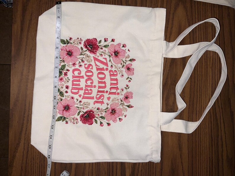 K&ouml;nnte beinhalten: Eine wei&szlig;e Canvas-Tragetasche mit zwei Griffen. Die Tasche hat ein florales Kranzdesign in Rosa und Rot, das den Text "anti Zionist social club" umgibt. Die Tasche ist etwa 43 cm lang.
