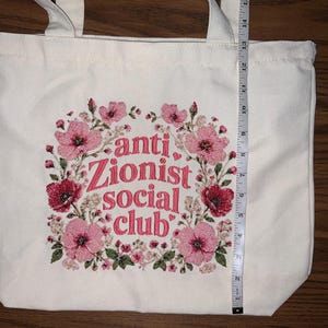 K&ouml;nnte beinhalten: Cremefarbene Tragetasche mit floralem Design und dem Text "anti Zionist social club" in Pink. Die Tasche ist etwa 35,5 cm hoch, mit einem floralen Kranzdesign in Pink und Rot.
