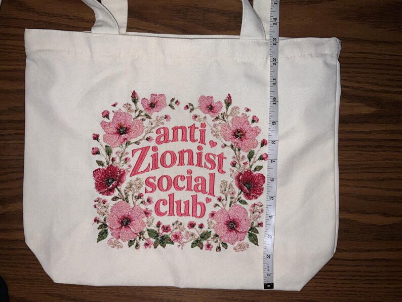 K&ouml;nnte beinhalten: Cremefarbene Tragetasche mit floralem Design und dem Text "anti Zionist social club" in Pink. Die Tasche ist etwa 35,5 cm hoch, mit einem floralen Kranzdesign in Pink und Rot.