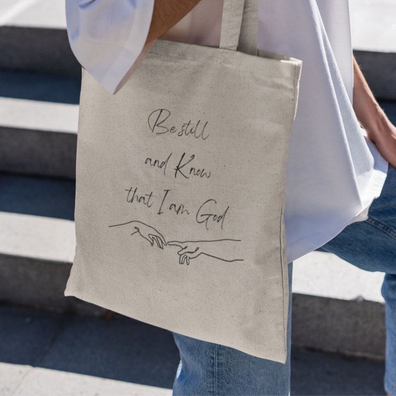 May include: A beige tote bag with black text and a line drawing of two hands. The text reads "Be still and know that I am God."
