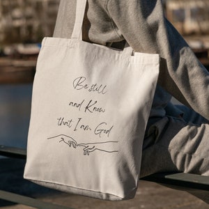 May include: White canvas tote bag with black text and a line drawing of two hands. The text reads "Be still and know that I am God."