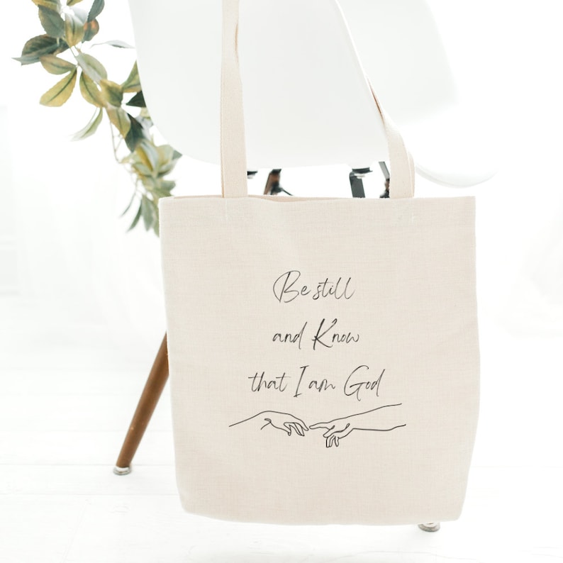 May include: A beige canvas tote bag featuring the text "Be still and know that I am God" and a simple line drawing of two hands reaching towards each other.