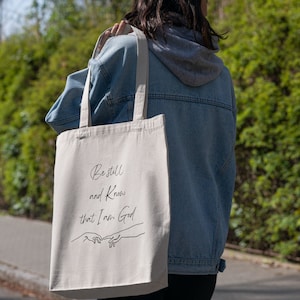 May include: A beige canvas tote bag with black text that reads "Be still and know that I am God". The text is in a cursive font. There are two hands drawn in black, one on top of the other, with the fingers touching. The bag is being held by a person wearing a blue denim jacket.