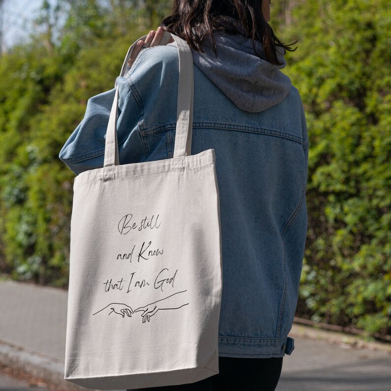 May include: A beige canvas tote bag with black text that reads "Be still and know that I am God". The text is in a cursive font. There are two hands drawn in black, one on top of the other, with the fingers touching. The bag is being held by a person wearing a blue denim jacket.