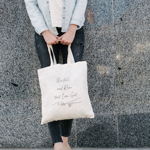 May include: A white canvas tote bag with black text that reads "Be still and know that I am God." The text is in a handwritten font. Two hands are drawn in black, reaching towards each other.