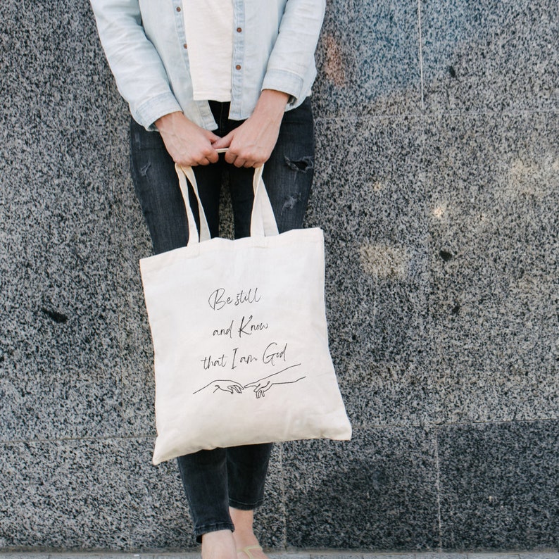 May include: A white canvas tote bag with black text that reads "Be still and know that I am God." The text is in a handwritten font. Two hands are drawn in black, reaching towards each other.