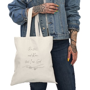 May include: A white canvas tote bag with black text that reads "Be still and know that I am God". The text is in a cursive font and is accompanied by a simple line drawing of two hands reaching towards each other.
